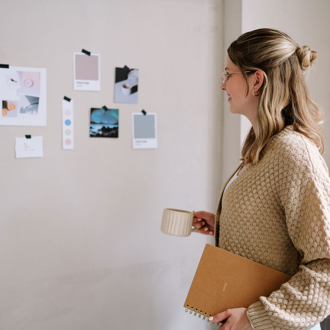 AuriStudio_ClaudiaSimchen-97_1zu1 Frau im beigem Cardigan schaut sich lächelnd ein Moodboard an einer Wand an